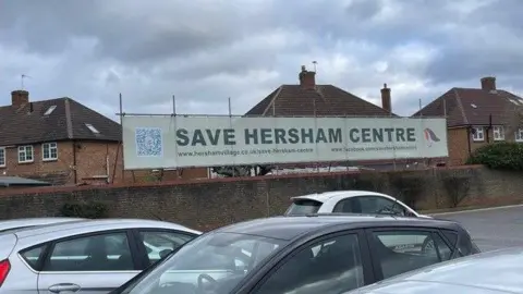 Hersham Centre Campaign A car park with a banner taped to a fence which reads "Save Hersham Centre"