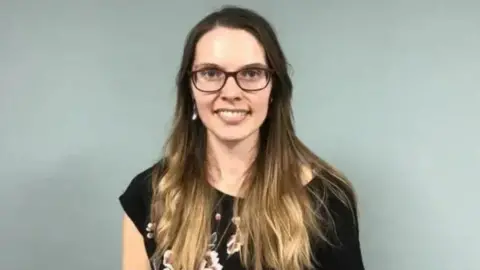 Joney Faragher looking straight ahead smiling. She has long brown hair and is wearing glasses and a black top.