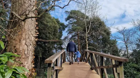 Glendypops/BBC Weather Watchers A man wearing a blue coat and with grey trousers, walks away from the camera over a wooden bridge, holding the hand of a child in a blue coat and multi-coloured trousers. There are trees either side of the bridge.