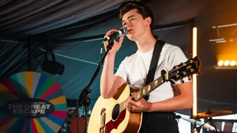 Getty Images Reay sings into a microphone on stage at a Brighton gig. He is wearing a white vest top tucked into belted black trousers and holds an acoustic guitar.