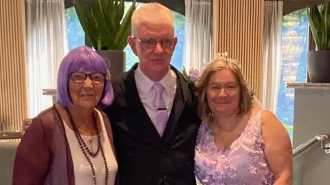 Marjorie Hayward Marjorie, John, and Tina standing together for a photo. Marjorie has purple hair and is smiling. John has short white hair, and Tina has shoulder length brown hair and is smiling.