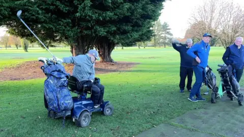 Terry Kirby with a golf club raised in mid-swing on a green fairway. He is sitting in a blue buggy and wearing a light-grey jacket, matching cap and dark-grey trousers. A large tree stands behind him and to his right are three fellow golfers, all dressed in blue.