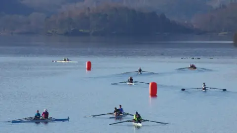 Simon Bamforth Direct shot of eight different boats in a lake being rowed. The water is clear. There are two orange buoys in the middle of the shot. 