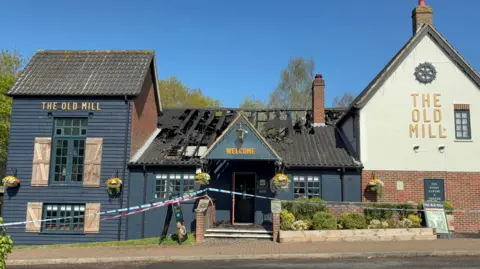Police tape runs across the entrance to a blue-coloured pub building, with a badly damaged roof caused by a fire in view on a sunny day