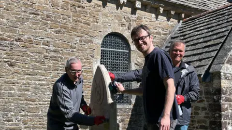 NRWGC Prof Ed Coss, Dr Zack White and CSM Alexander McMillan turn to smile at the camera while each of them is working on different parts of the gravestone