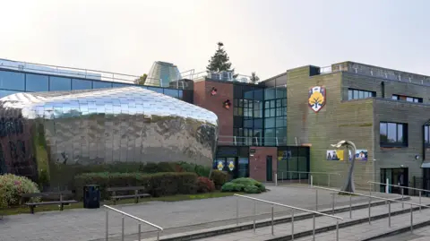 general view of St Regis CE Academy in Wolverhampton - rectangular buildings with a large silver round section at the front near the entrance. A logo can be spotted on the building on the right