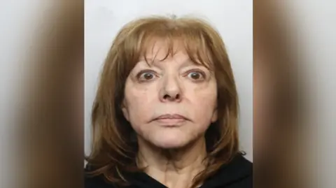 A woman with shoulder-length dark blonde hair is looking at the camera in a police custody shot. There is a Greater Manchester Police logo in the top left hand corner.