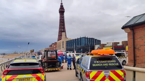 Several HM Coastguard vehicles parked on Blackpool promenade during Blackpool Airshow with coastguard officers nearby in blue uniforms. Blackpool Tower is in the background and the sea is to the left.