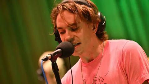 Sports Team in session for BBC 6 Music. The photo shows vocalist Alex Rice at Maida Vale Studios, singing into a microphone while wearing headphones.