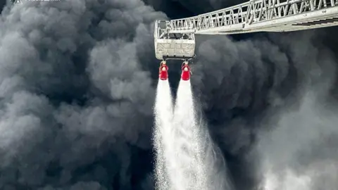 DSNS Thick dark grey smoke billows from the Brody oil hub as firefighters tackle an unseen fire with two hoses from an extended ladder.