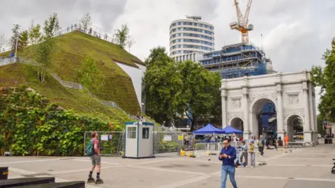 Getty Images A large grass mound sits beside Marble Arch as a pedestrian and a man on rollerblades pass by. The mound is taller than the arch and has grass and plants on it. 