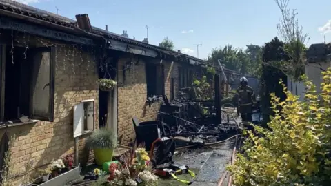 Terraced bungalows that have been badly burned. There is rubble on the pavement and the buildings have turned black due to the fire, which is now out. There are bushes and pot plants in the foreground and fire crews working in the background.