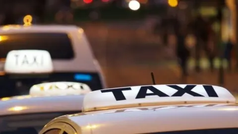 A stock image of the tops of a row of taxis stretching into the distance. The taxi signs on the roofs are lit up.