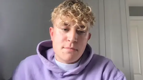 Lewis Rollins has dyed blonde hair and a purple jumper on.