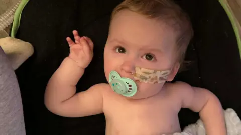A baby is lying on a black mat with a blanket wrapped around his stomach. A blue dummy is in his mouth and a tube is attached into his nose.