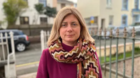 A woman wearing a pink jumper and a scarf looks into a camera. She is standing outdoors. A road and a metal fence are behind her. Cars are parked on the road. Buildings are on the road behind. 