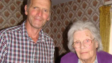 Rod Smart is stood next to his mother, Diana. They both have a small smile on their face and are looking at the camera. Rod is wearing a checkered shirt and Diana has a purple cardigan and pale coloured glasses. 