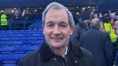 George King/BBC George Burley smiles at the camera as he stands within Portman Road stadium close to the blue fan seating area. He has short grey hair and wears a black coat. People can be seen behind him to the right talking with each other. 
