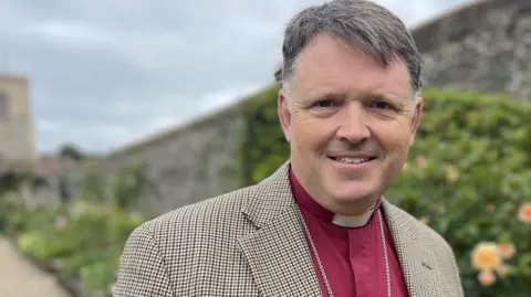 Bishop Graham in a pink clerical shirt, white dog collar and beige or brown jacket with small checks, standing in the Bishop's Garden. The background is blurred, with a flower border and a flint wall behind him.