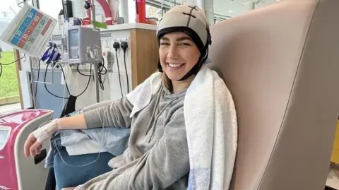 Kim Christon Kim Christon, in a hospital chair, having chemotherapy, wearing a cold cap, with a towel round her shoulders, wearing a grey top. She is smiling and hospital equipment is to her left. 