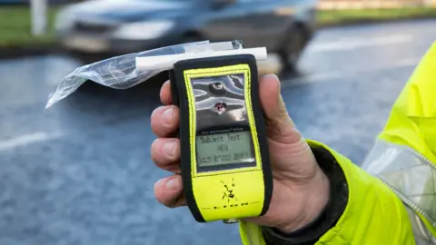 A person holding a breathalyzer device with a plastic-covered mouthpiece. The device's screen displays the text: 'Subject Test: NEW WAIT BUTTON READY'.