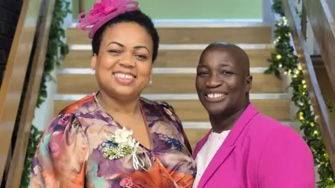 Cheryl Pierre is on the left and is wearing a floral patterned dress and a pink fascinator on the left. Letitia Cuney is on the right and is wearing a hot pink suit jacket and a white T-shirt. They are stood next to each other in front of wooden stairs.