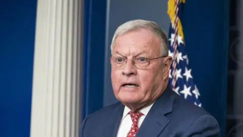 Getty Images Keith Kellog, a grey-haired man wearing glasses, stands at the White House briefing room podium while wearing a navy suit, white shirt and red tie.