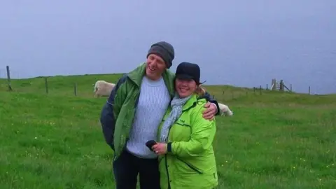Sarah Thompson A man and woman smiling on a green field, the man wears a grey hat, blue striped jumper and green coat and the woman a black hair, gray scarf and green coat. 