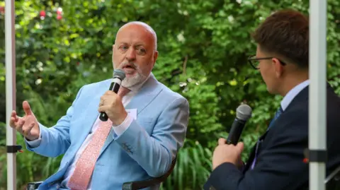 ED CRISPIN/DUKE OF EDINBURGH'S AWARD Bill Bailey with short light-coloured hair and beard, wearing a light blue jacket and sand-coloured tie. He is holding a microphone with one hand and holding the other hand out in a gesture. He is sitting with a man with short dark hair and glasses, wearing a dark jacket and holding a microphone. They are sitting in front of green foliage.