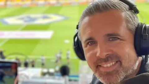 Ryan is in the ground with headphones on ahead of a match. He has grey hair and a beard and us turning around form his desk to have the photo taken. The pitch can be seen in the background.