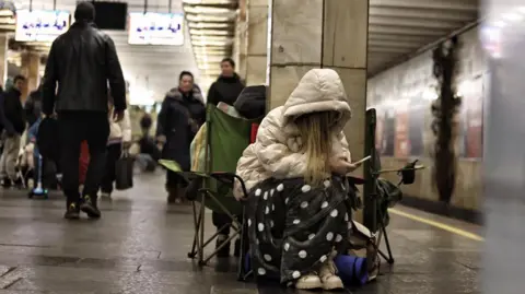 Getty Images A woman sitting on a chair with the hood of her cream-coloured coat covering her face and a spotted blanket while she is on her mobile phone. Behind her, there are multiple people walking in the subway in Kyiv, where they hid during a Russian attack on 28 November, 2024