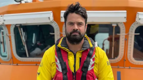 Simon Jones/BBC Dan Senkler - A man with black hair, black beard and black mustache. He wears a yellow RNLI suit and a red survival jacket. The photo was taken from a boat in the sea.