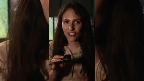  A woman with long dark hair holds a tiny microphone in front of her.