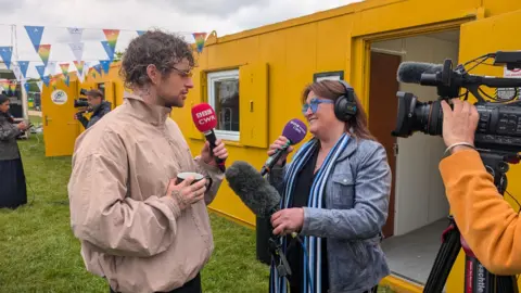 Tom Grennan is wearing a light brown jacket and holding a red BBC CWR microphone. He is speaking to radio presenter Lorna Bailey, who is in a denim jacket and holding two microphones. there is a TV camera on the right and yellow demountables and white marquees in background