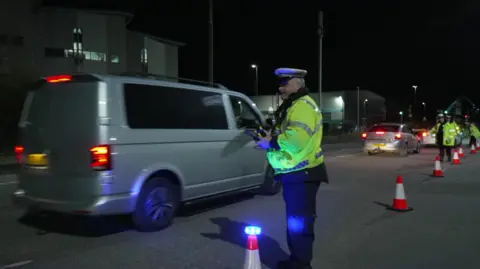A police officer stands by a row of cones at the ferry port as a cars file past.
