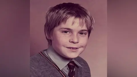 Family photo An old school photograph showing a young boy with freckles. He is wearing a grey shirt and a black and gold tie under a grey V-neck jumper.