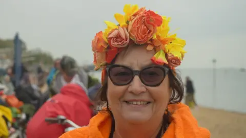 A smiling Marcelle Baum looks directly at the camera as she is photographed outside at the beach. She is wearing sunglasses and a flowery headband and there are spectators sitting on the beach behind her. She looks like she has just been for a swim and is wearing an orange towel robe