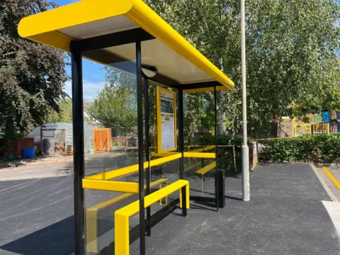 A yellow bus stop with glass sits on a tarmac pavement and a lamppost. There are trees in the background and a greenhouse in the distance.