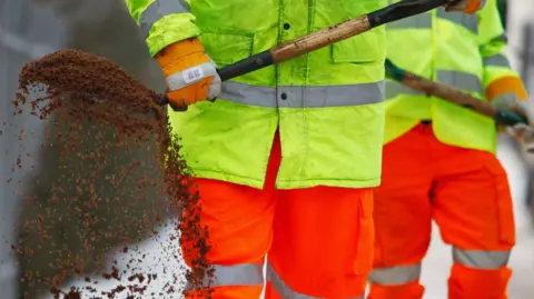 Getty Images Two workers wearing high visibility jackets and trousers use shovels to spread grit on a pavement.