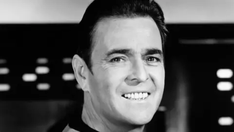 Getty Images A black-and-white close-up publicity shot of a man in his mid-40s with short, dark hair, smiling looking away to the left of frame with his eyes, although his face is facing the right of frame. He wears a top which looks grey in the photo, with a black collar.