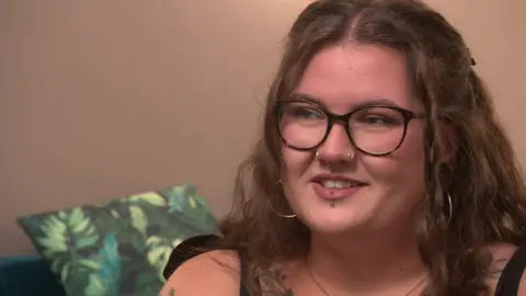 Ez speaks off camera. She has light brown wavy hair, a lip stud, nose rings, and hooped earrings. She also has black rimmed glasses. 