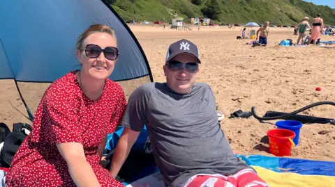 A man and a woman sit on Exmouth beach on a very sunny day. They are in front of a small tent to shade them from the sun. The sand is golden and there are people in swimming costumes in the background.