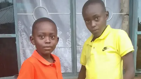 The sons of Caroline Odor Caroline Odor-Daniel, 12-year-old on the right in a yellow shirt and on the left, Ilya, nine-year-old. They stand outside in front of a window with a white curtain.