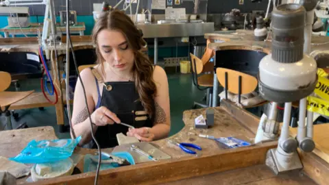 A woman sits a at table making jewellery. She has an apron on and long brown hair tied to one side. She is in a workshop.