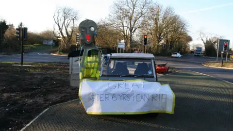 Michael Maxwell A strange alien-like figure with a high-vis jacket and attached to a car and a sign which says 'Great, After 6 Years I can RIP'