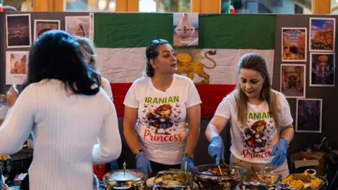 Ahassona A food stall at a previous Refugee Week is pictured. The stall in the shot is an Iranian food stall which is staffed by two women in white t-shirts bearing the legend Iranian Princess. A third person, with back to camera, is standing over the stall.