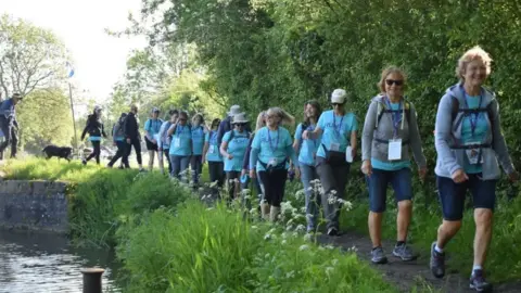 RUHX A group of walkers in the same turquoise t-shirts with RUH written on the front and walking gear and rucksacks. They are walking in single file along a narrow footpath in the countryside beside a river to the left of the image and a hedge of trees on the right