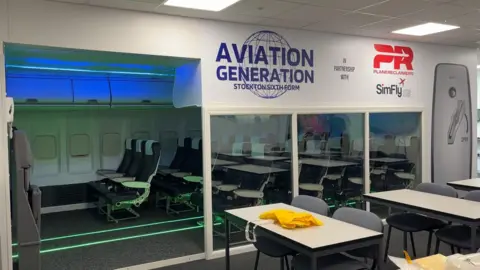 A classroom with tables and chairs and a replica boeing cabin behind with an aisle of seats with plane windows. Above the entrance can be read: "Aviation Generation Stockton Sixth Form.