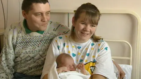 A couple in hospital with their baby. The father is on the left and he has short cropped dark hair and a patterned jumper. The mother is in a nightie with blue and yellow hearts on it and is holding a small child in her arms.