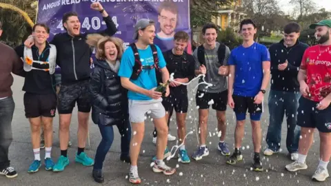 Angus Read A man with a blue tshirt, grey cap and grey shorts with a crowd of people, shaking a bottle of champagne